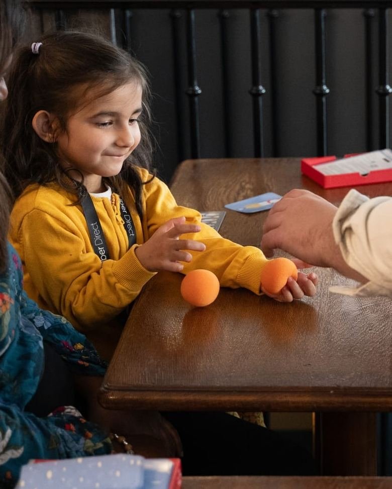 Pendant le spectacle, une petite fille en pull jaune sourit devant deux balles orange posées sur la table